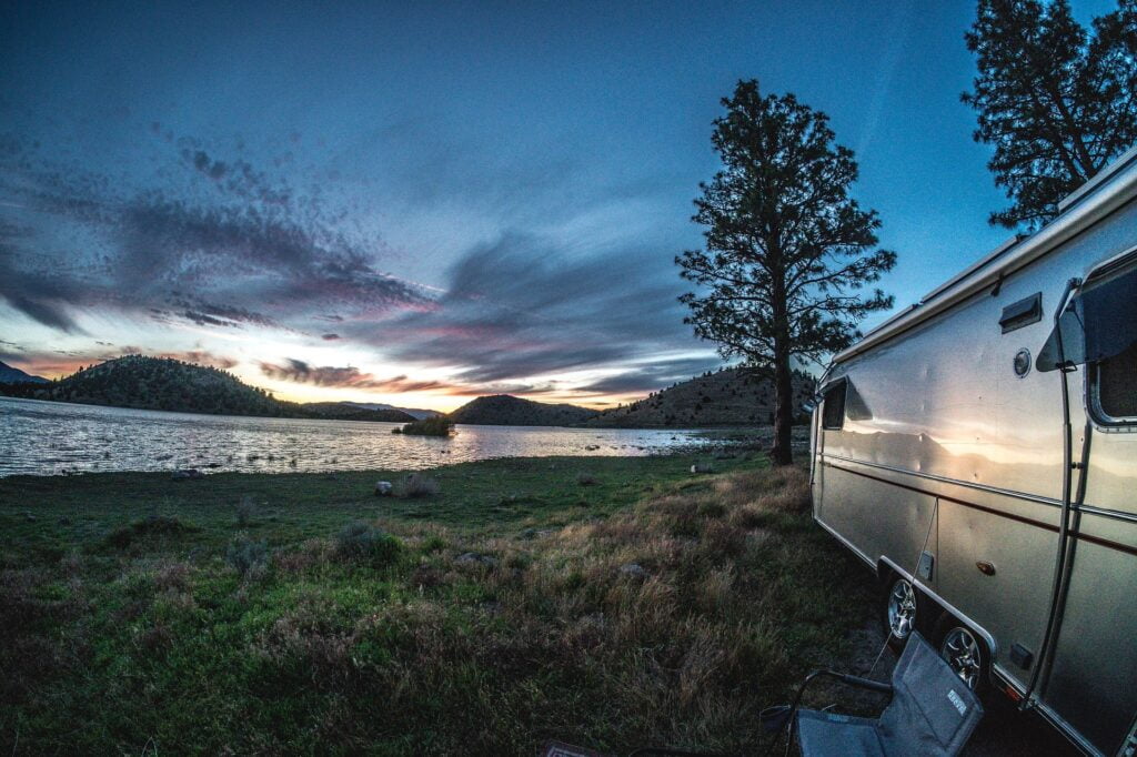 Metal-sided RV with folding chair by a lakeshore at sunset