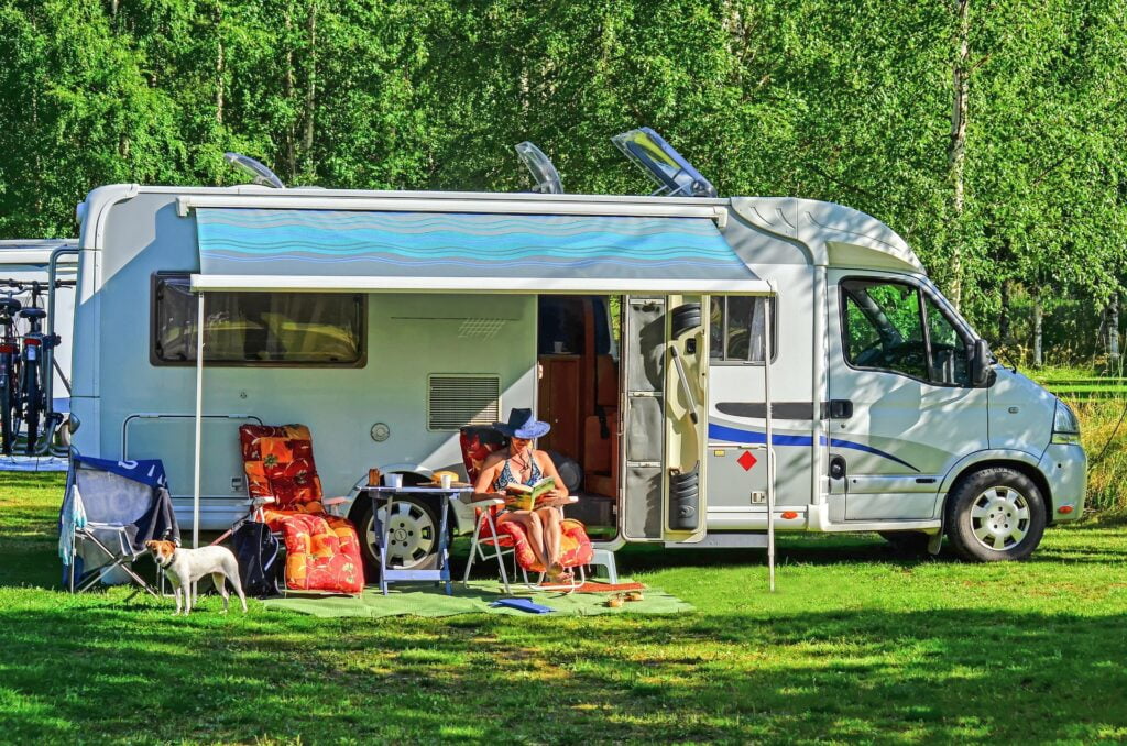 Woman in hat reading a book and sitting under RV awning on the grass with a dog