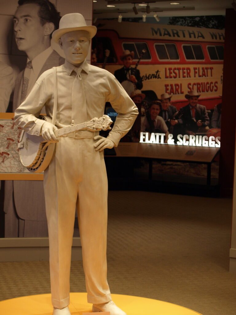 A statue of Earl Scruggs standing in front of a bus inside the Earl Scruggs Museum.