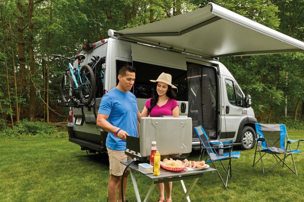 A couple BBQing in front of their Thor Rize RV with an awning. 