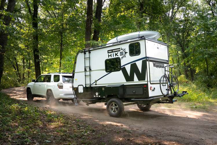 A white pickup truck pulling a Winnebago Hike RV on a dirt road surrounded by green trees.