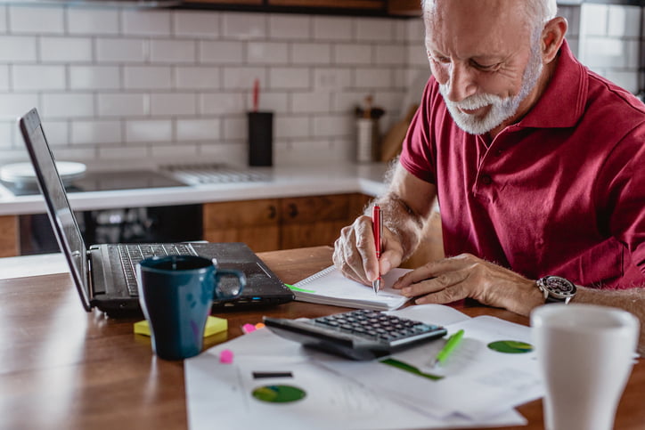 Senior man is sitting at the table, using a laptop to plan a big purchase and writing something on the paper.