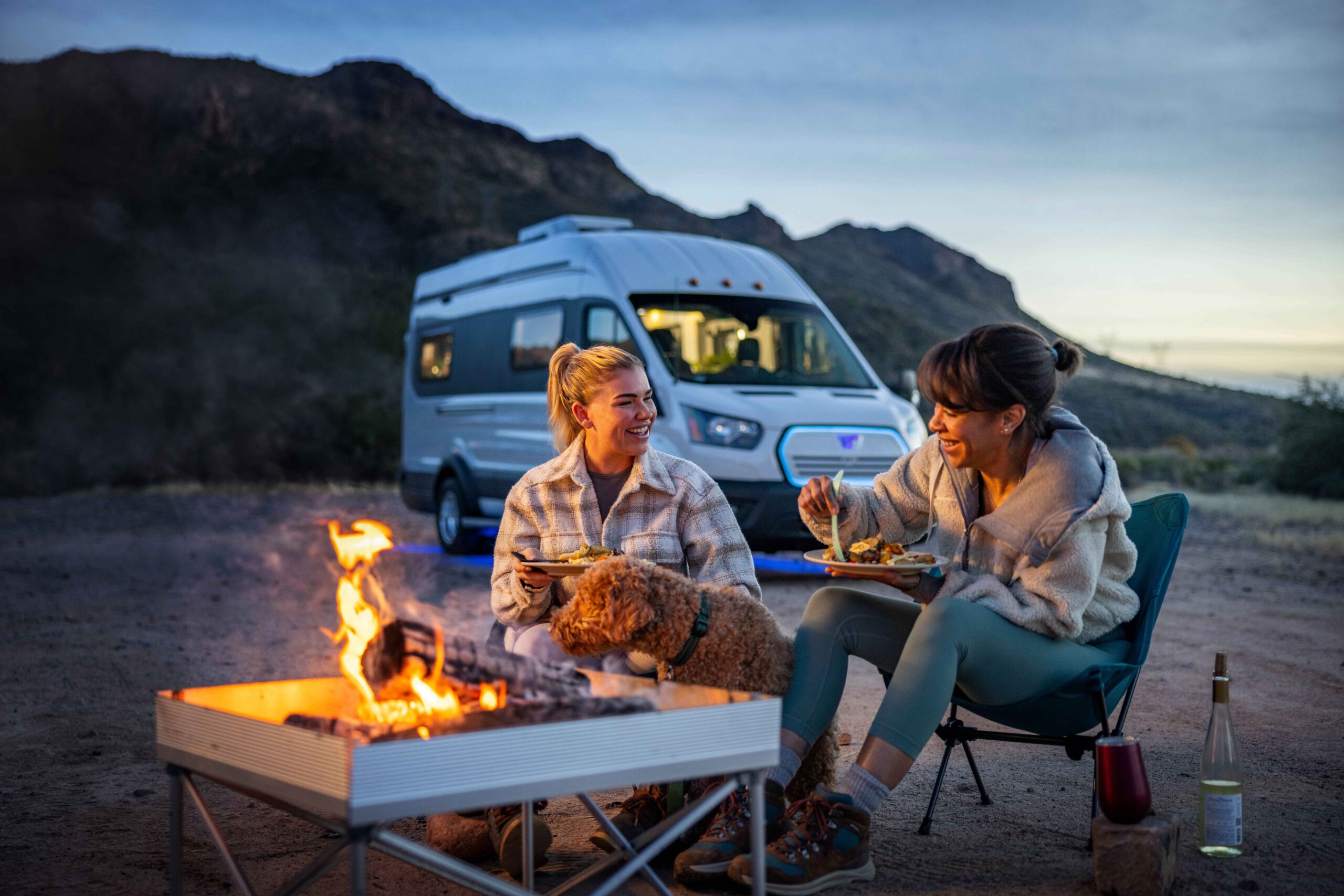 Two women sitting outdoors eating a meal beside their fire pit. A dog is sitting next to them. A Winnebgo e-RV is parked behind them.