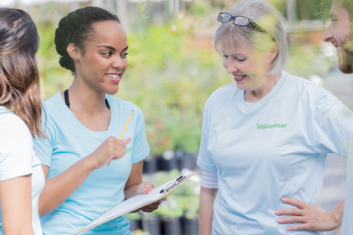 A cheerful young woman stands in a plant nursery with a group of volunteers and discusses the plan for their day.