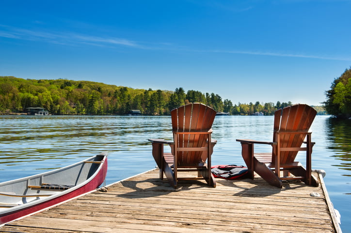 Two Muskoka chairs on a wooden dock overlooking the blue water of a lake in Muskoka, Ontario Canada. A red canoe is tied to the pier and life jackets are visible near the chairs.