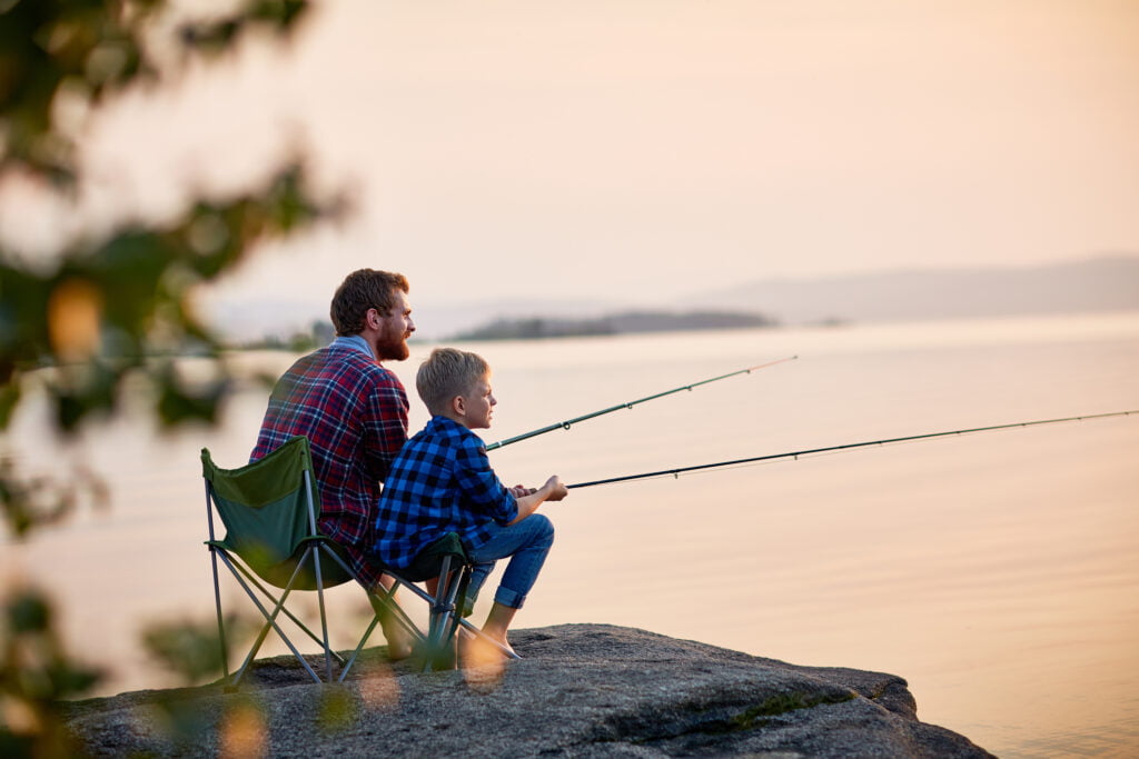 Side view portrait of dad and son sitting outdoors together on rocks fishing with rods in calm lake waters with landscape of setting sun, both wearing plaid shirts, shot from behind tree