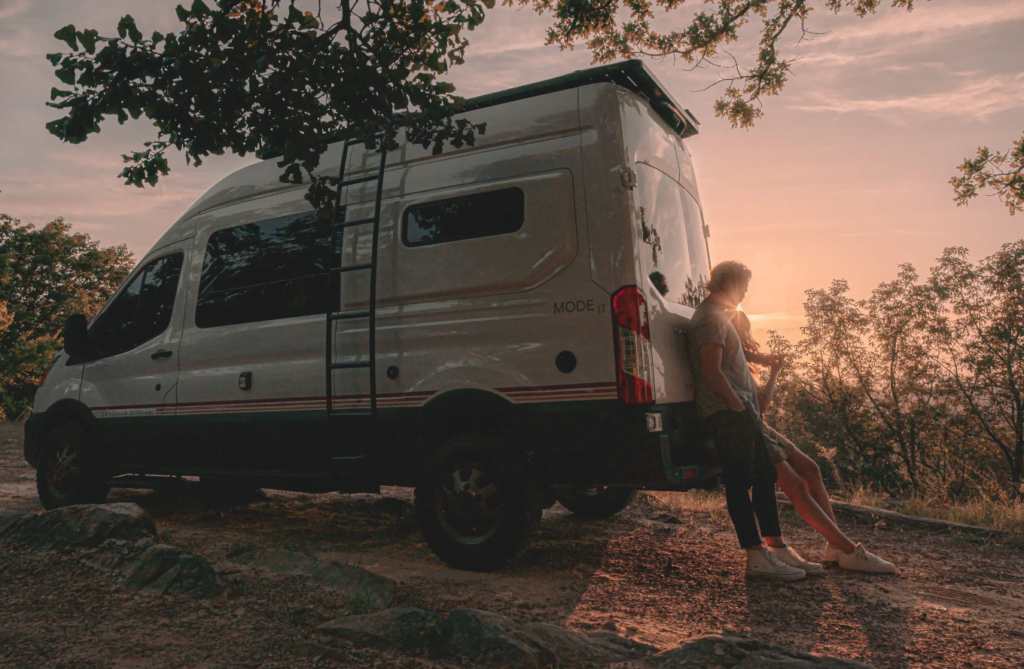 A man and woman standing outside a Storyteller Overland Mode LT Class B motorhome on a dirt road.