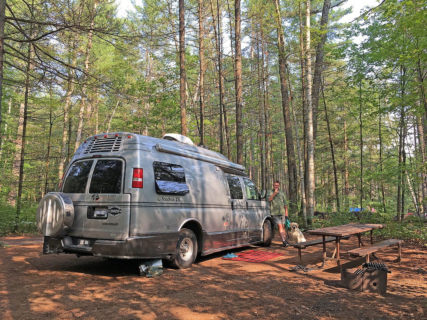 A Man and his dog on a leash leaning on a camper van parked at a forested RV site in Bon Echo Provincial Park, in the Ottawa valley.
