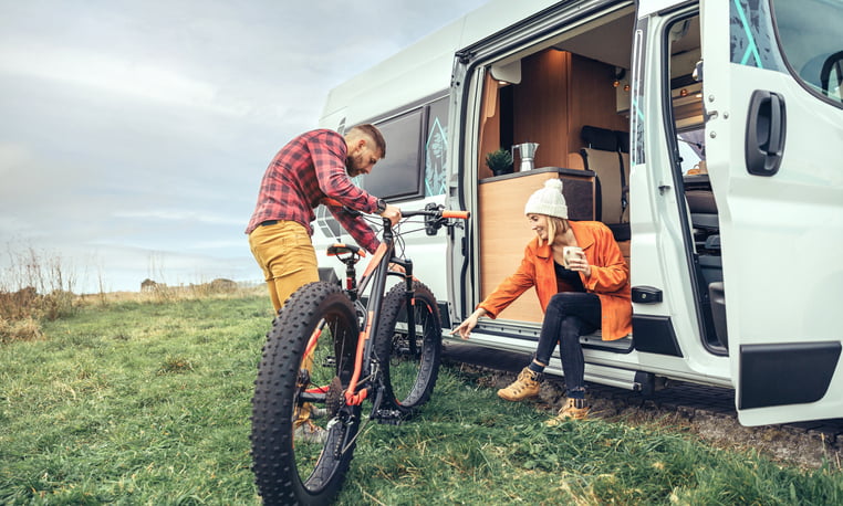 Woman drinking coffee sitting at the door of a class b motorhome and pointing at a bike wheel held by a man.
