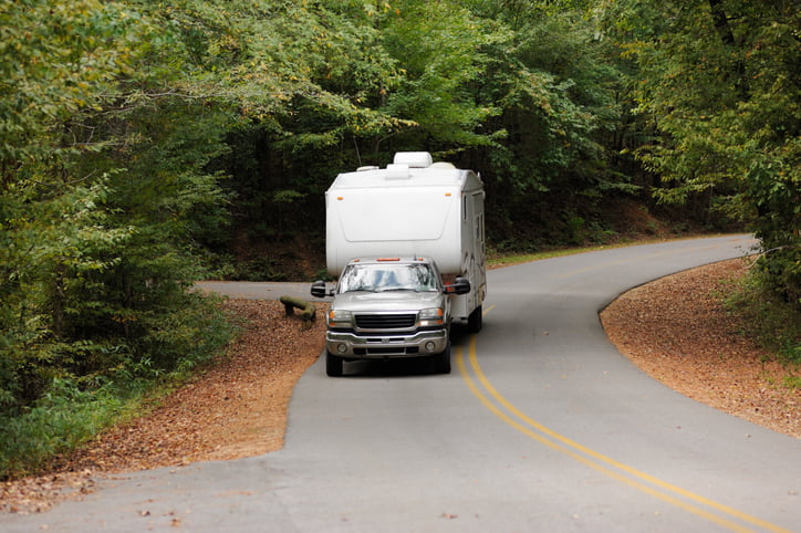 A silver pickup truck is pulling a camper trailer down a quiet treelined road.