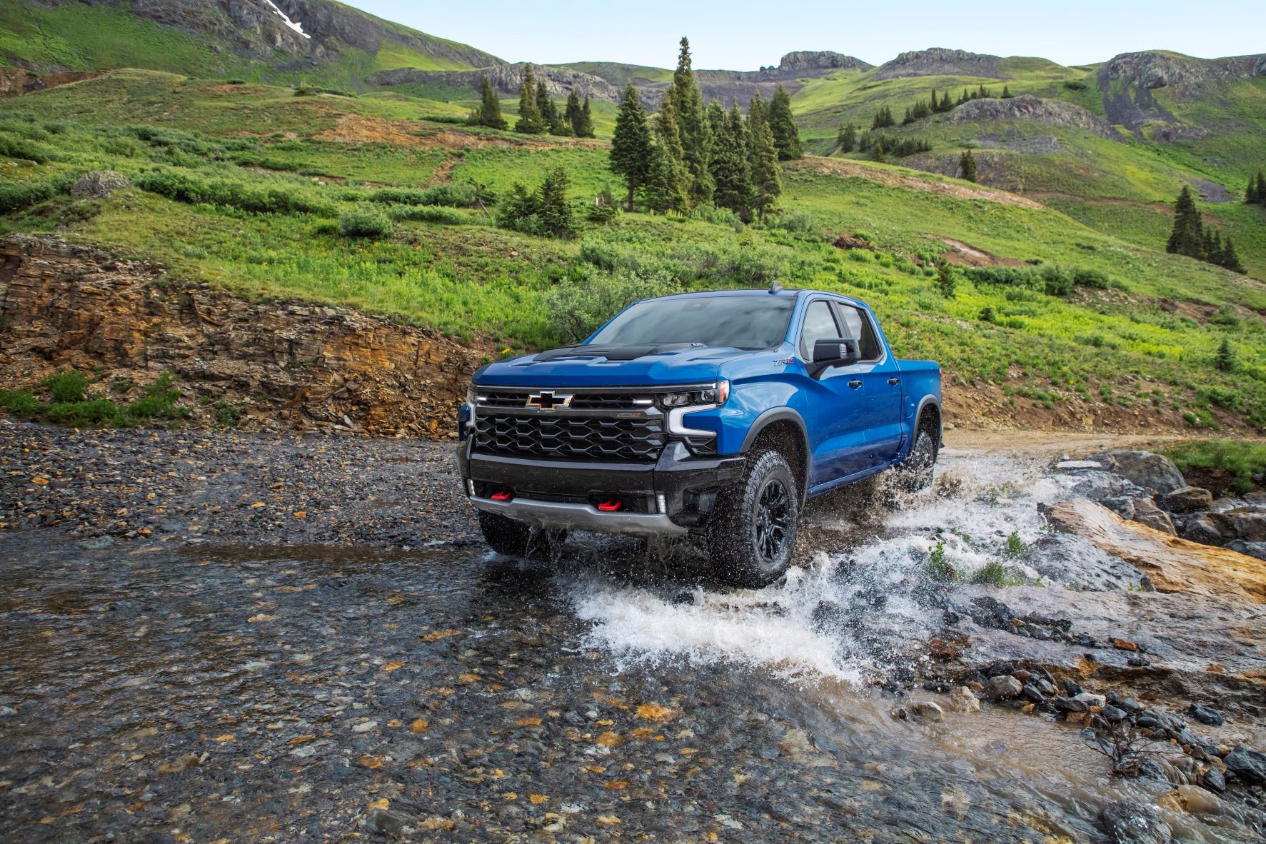 A blue 2022 Chevrolet Silverado ZR2 parked in the shallow moving water of a river. Behind the truck are low, rocky, hills spotted with grass and clusters of evergreen trees.
