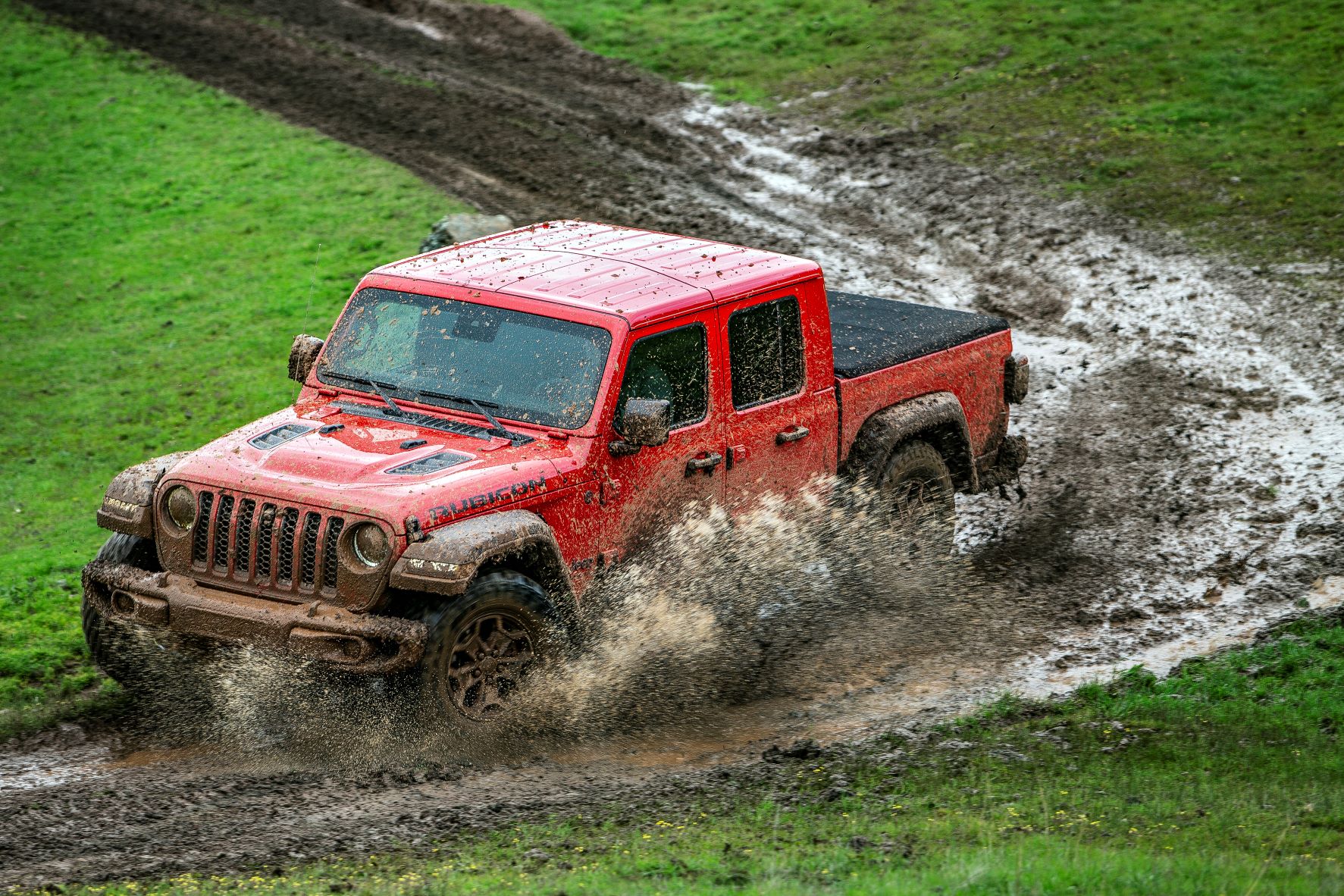 A red Jeep Gladiator drives downhill through the mud, sending a spray of mud towards the viewer. 