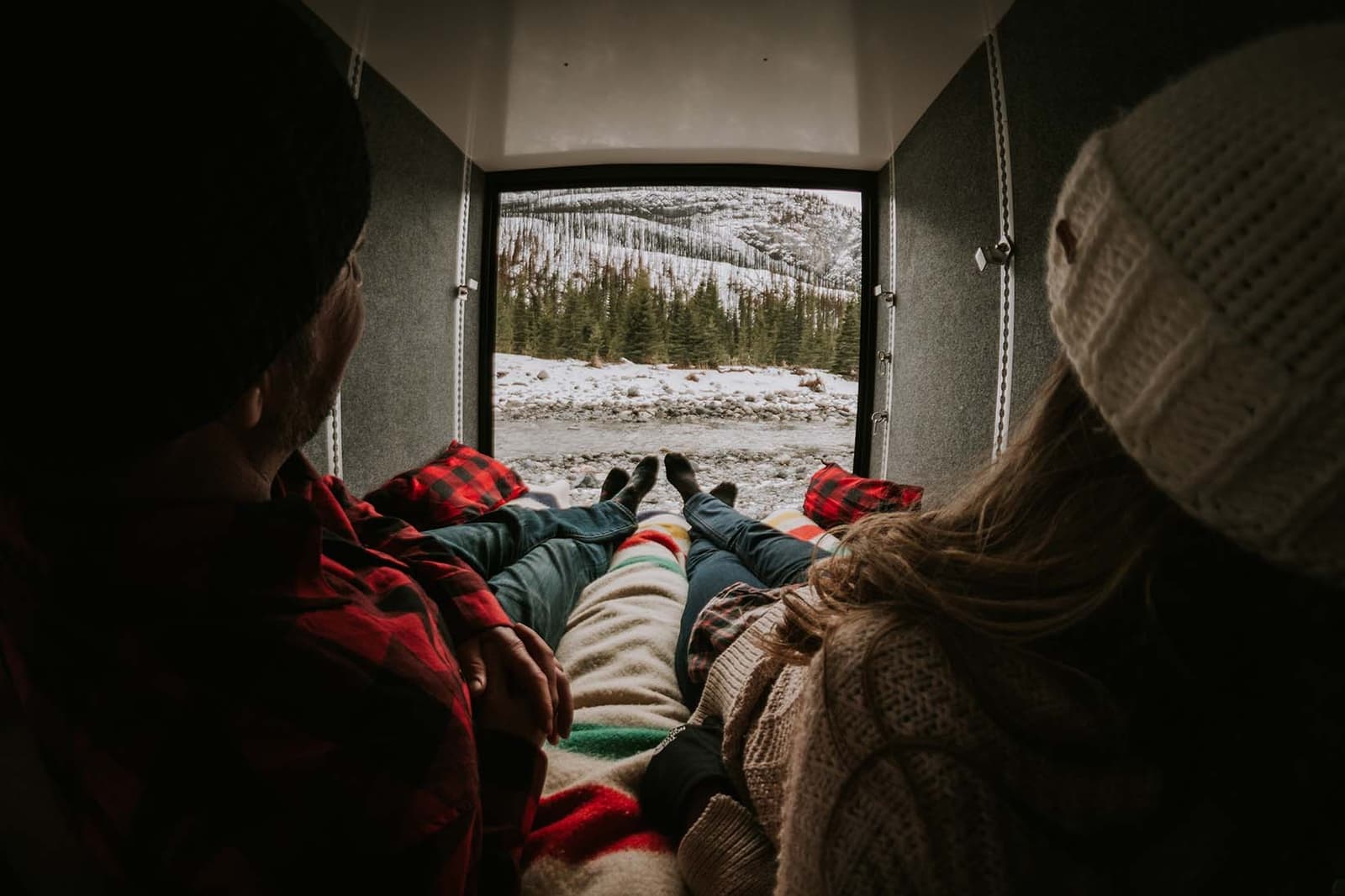 A man and woman wearing toques are reclined side-by-side inside their overland trailer. They are looking out the open door at a spectacular rocky winter landscape. An icy river separates them from evergreens and nearby mountains.