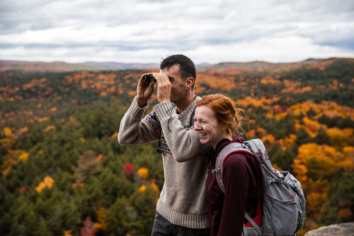 A couple in their 20s take a break from their hike to take in the stunning beauty of the Fall leaves from their rocky vantage point. The landscape is peppered with shades of orange, red, and the green of evergreens.