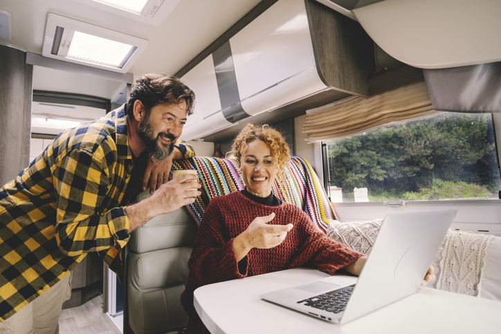 A smiling Black woman with lightened, curly, hair sitting in her RV. Her partner, a man with dark hair and a greying beard, is leaning over her shoulder, looking at their laptop.