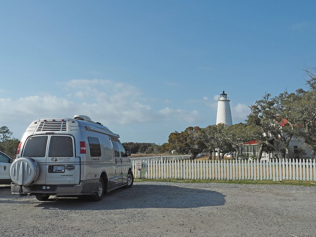 A camper drives up the road towards a white lighthouse on Ocracoke Island.