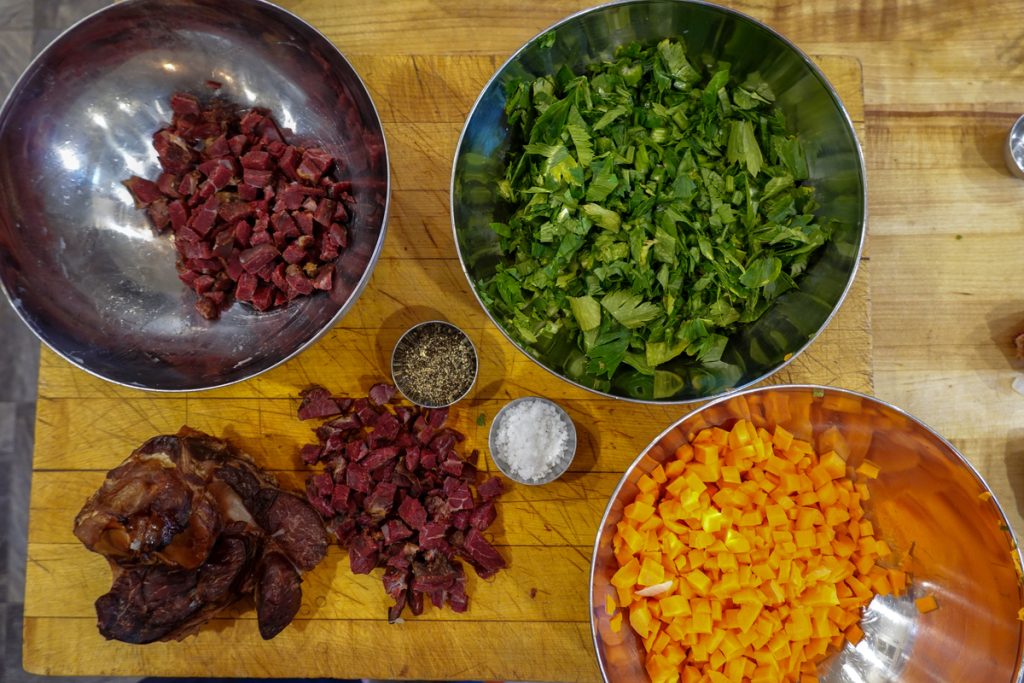 Three stainless steel bowls sit on a wooden cutting board. One has cured meat in it, one chopped parsley, and the last has carrots, peeled and diced, in it. 