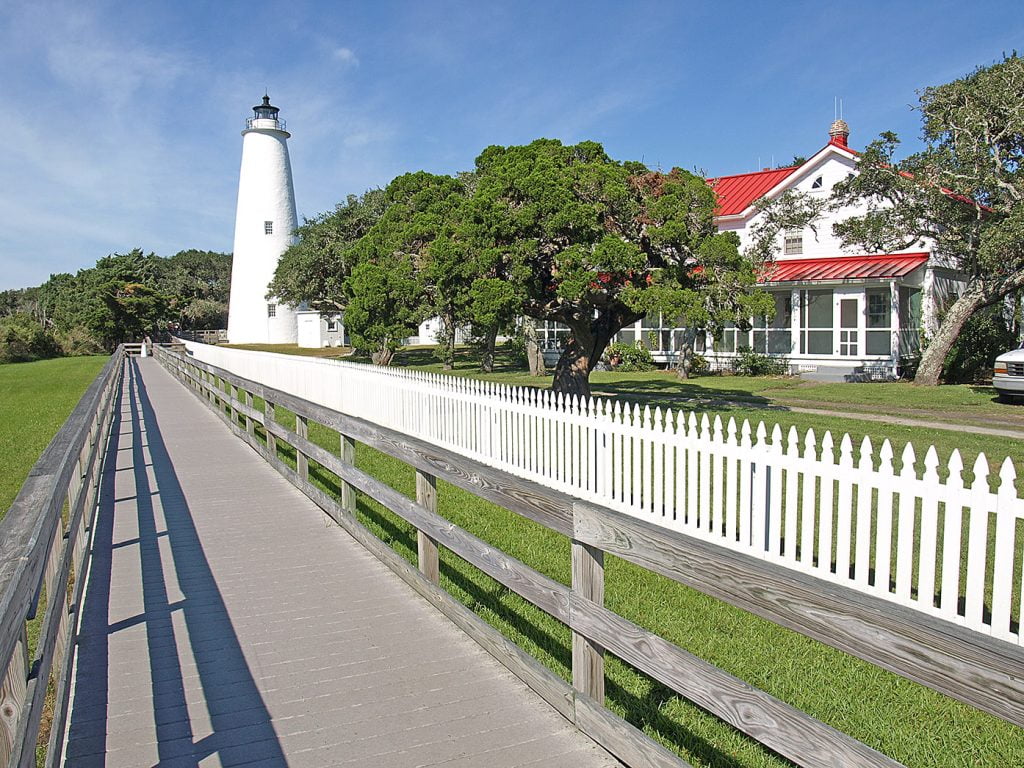 A walking path lined with cedar rail fencing runs parallel to a white picket fence. The path leads to an old white lighthouse standing twice as tall as the trees around it.  