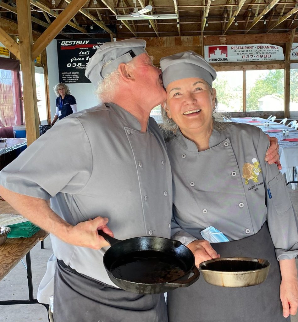 Gail and Martin Aller-Stead, a white man and woman both with grey-white hair, posing for a photo. They are dressed in grey chef’s attire. Gail is looking at the camera. Martin, with his arm around Gail, is giving her a kiss on the cheek. 