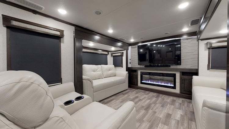 The living area inside the 2023 Crossroads Redwood 4200FL. Leather recliners and loveseats are in a “u” formation around a gas fireplace and a large television mounted to the wall.