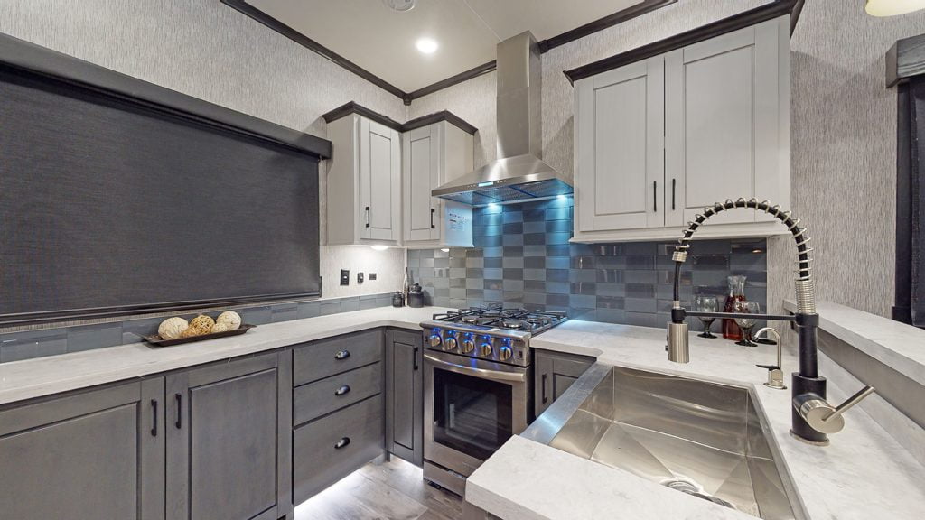 A stunning modern RV kitchen with ample counter and cupboard space. This kitchen features a stainless steel four-burner gas stove, range hood, and sink with an extendible faucet.