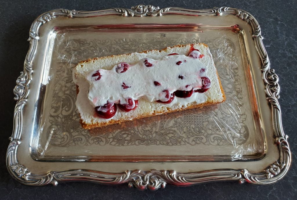 A piece of store-bought pound cake, sliced lengthwise, with a layer of whipped cream, a layer of cherry pie filling, and another layer of whipped cream spread over it. 