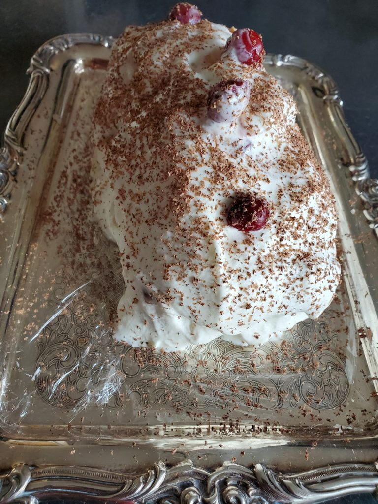 A close-up of a Yule log covered in whipped cream with cherries on top and grated dark chocolate on top, presented on a silver serving tray.