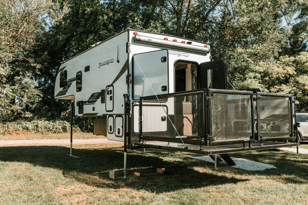 A Palomino Backpack Truck, Camper 2912, is parked on a lot surrounded by evergreen trees on a sunny day. Its fold-down porch is lowered, with an open backdoor and outdoor kitchen doors.