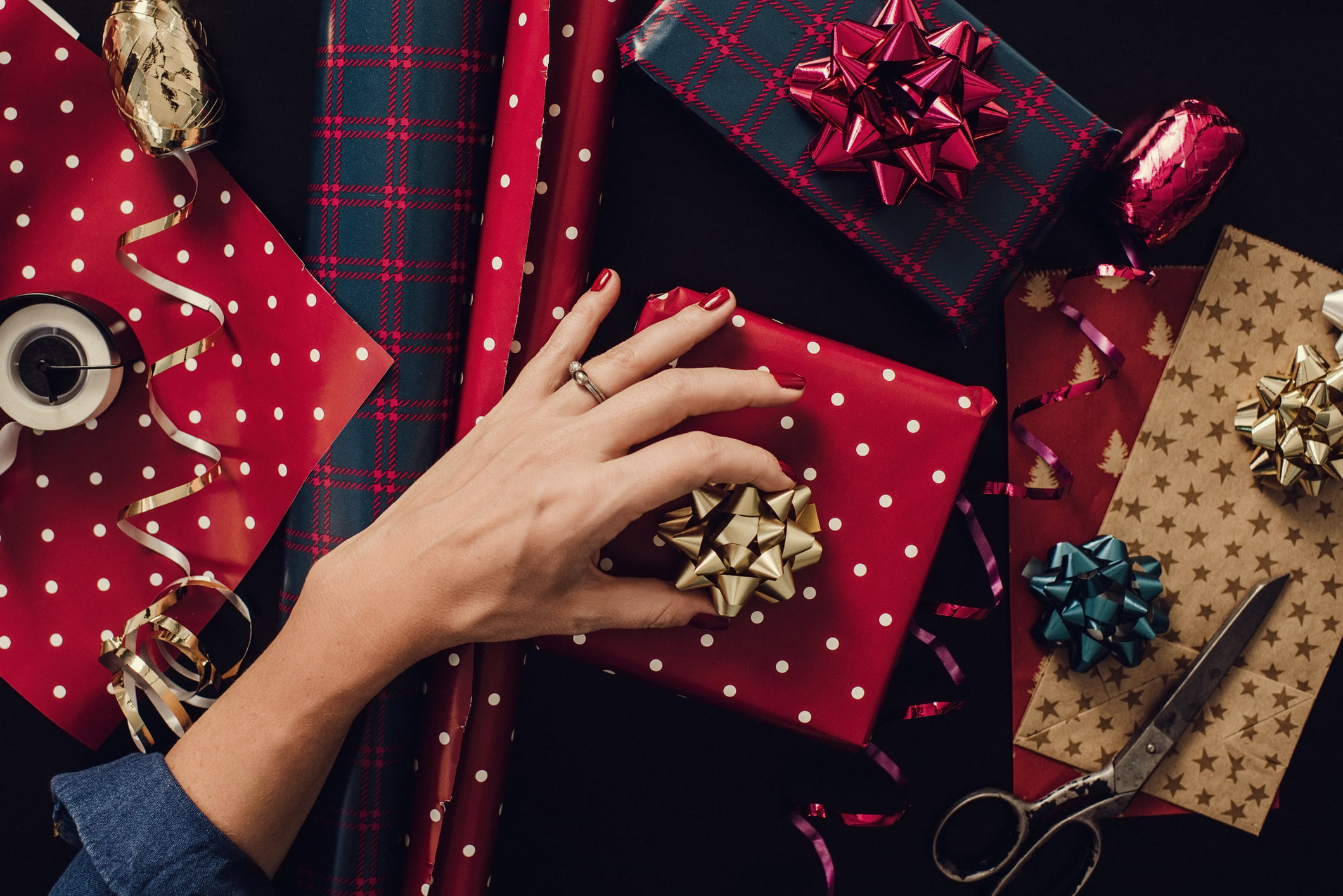 A hand with red painted nails and an engagement ring on its finger is placing a gold bow on a package wrapped in red paper with white polka-dots. Around the gift are rolls of holiday wrapping paper, ribbons, bows, and a pair of scissors.
