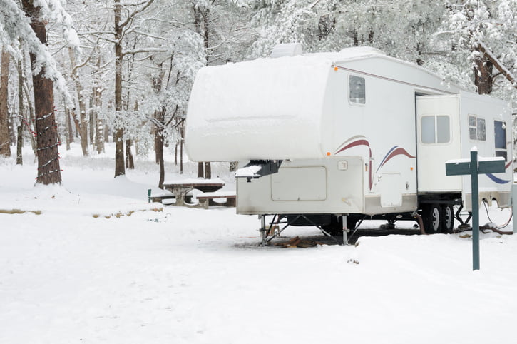 An RV is parked on a treed lot in the winter; there’s a cover of snow over everything.