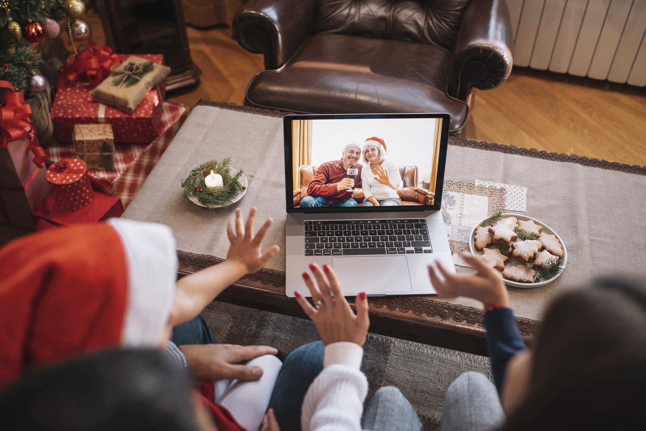 Two adults and a toddler sitting with their backs to the camera in a living room decorated for Christmas. They’re looking at an open laptop on the coffee table in front of them. Displayed on the screen are a man and a woman in their late 50s, sitting close together on a love seat. The man is smiling and holding a glass of red wine while the woman, wearing a Santa hat, waves.
