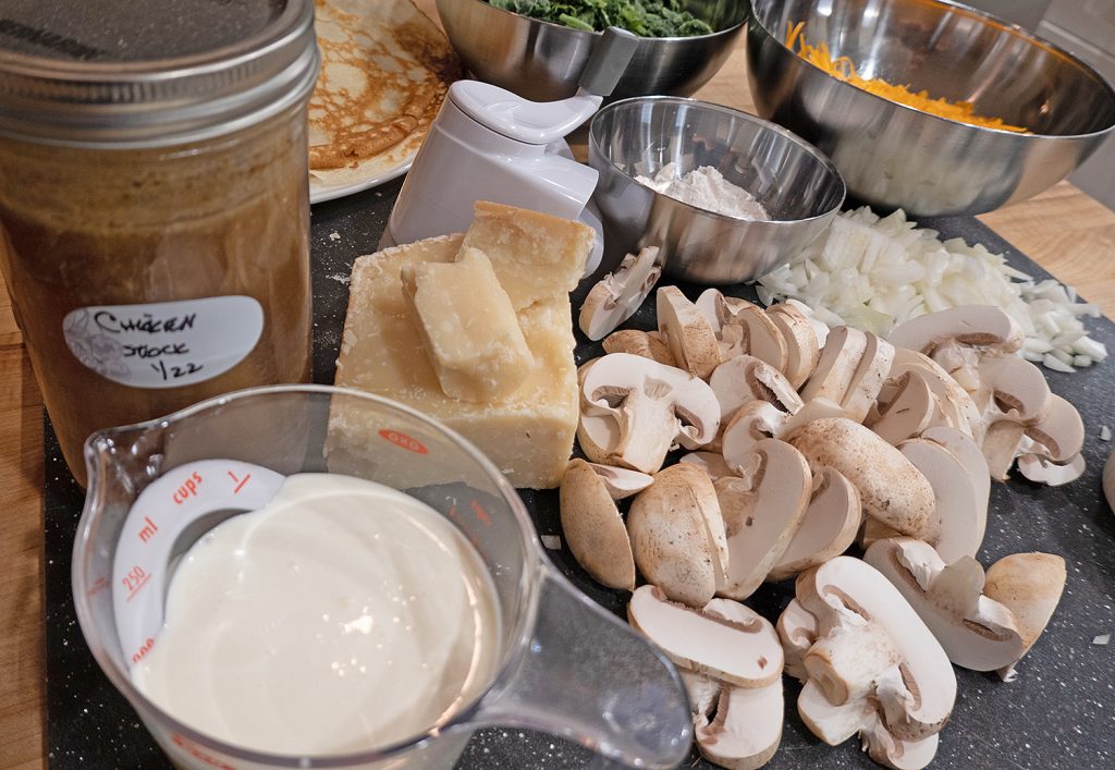 Ingredients for Spinach and Cheddar Crepes with Mushrooms, laid out on a slate cutting board including homemade chicken stock in a mason jar, a measuring cup with whipping cream in it, a block of parmesan cheese, sliced mushrooms, dices onion, a plate of pre-made crepes, a small stainless steel bowl half full of flour, and two large stainless steel bowls containing spinach and grated cheddar cheese.