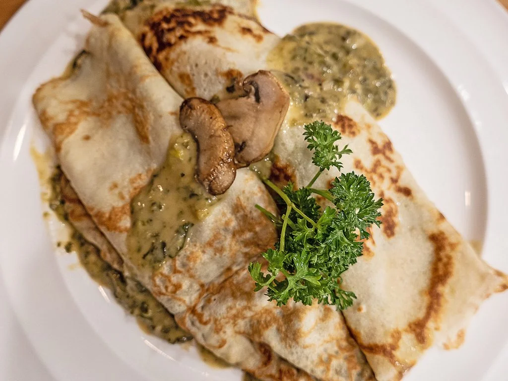 Two rolled crepes, topped with mushroom crem, two slices of browned mushroom, and parsley plated on a white dish.