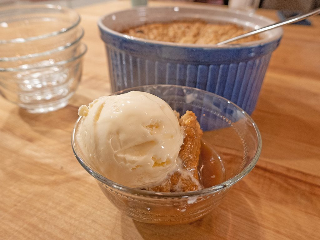 A small glass desert bowl with warm caramel pudding and a scoop of vanilla ice cream on top. Small trails of ice cream melt down the side of the pudding. Behind the desert dish is the baking dish with the rest of the pudding inside.