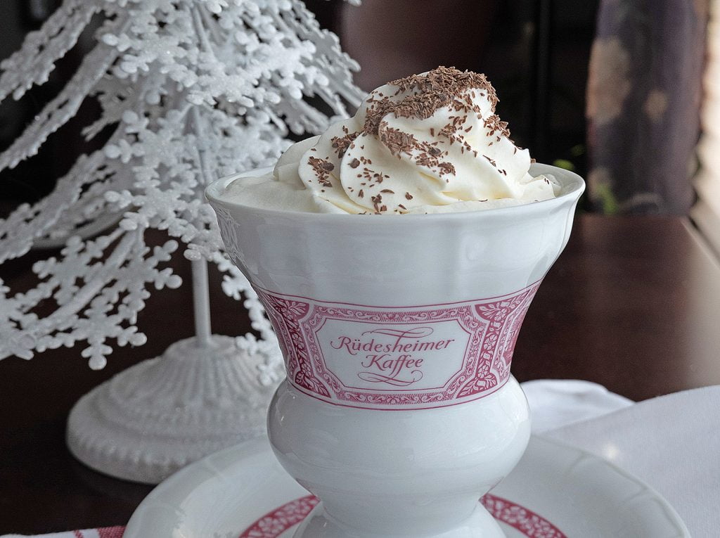 A tall, decorative, coffee mug sitting on a matching saucer. A swirl of whipped cream garnished with grated chocolate tops the hot chocolate in the mug. Behind the mug is a small, white, holiday tree adorned with glitter.