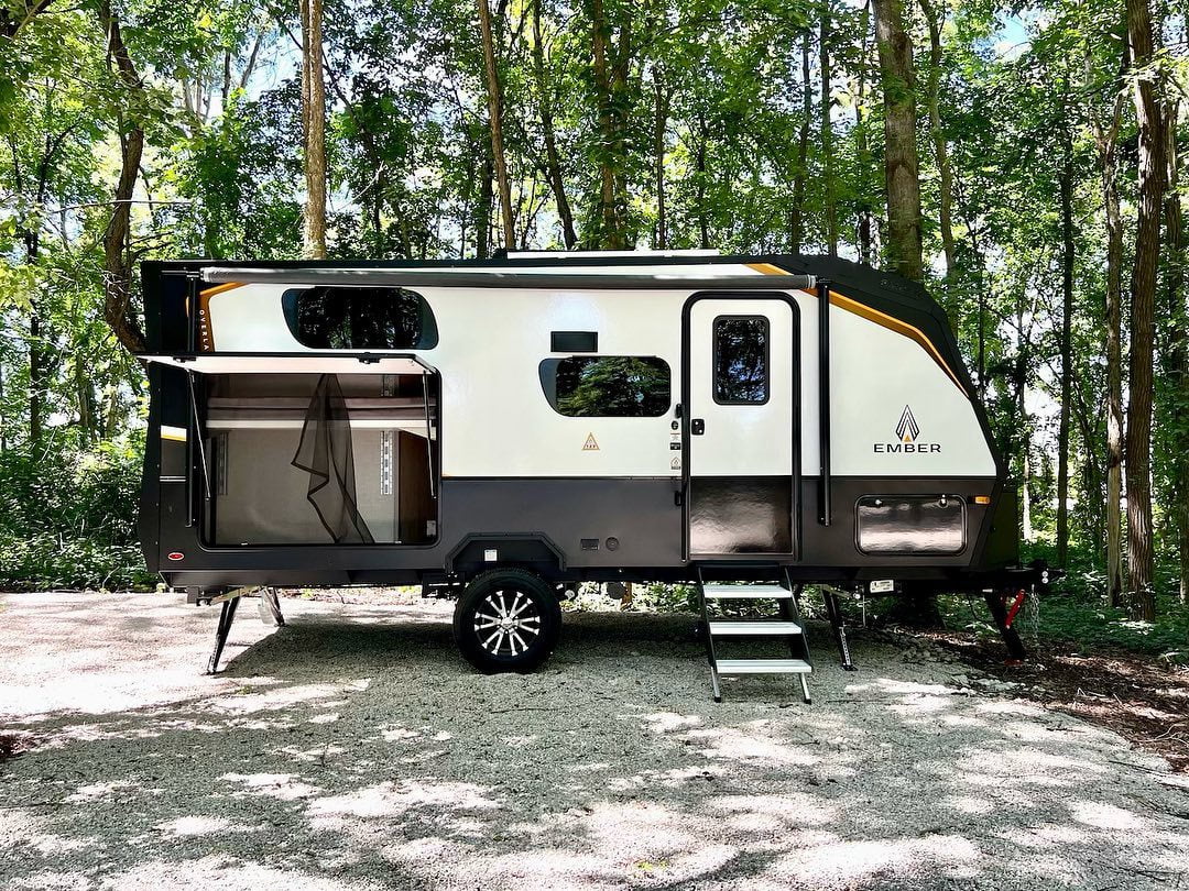 An Ember overland trailer, parked on a graveled campsite, surrounded by trees. The side pop-out is open and the steps to the front door are in place. 