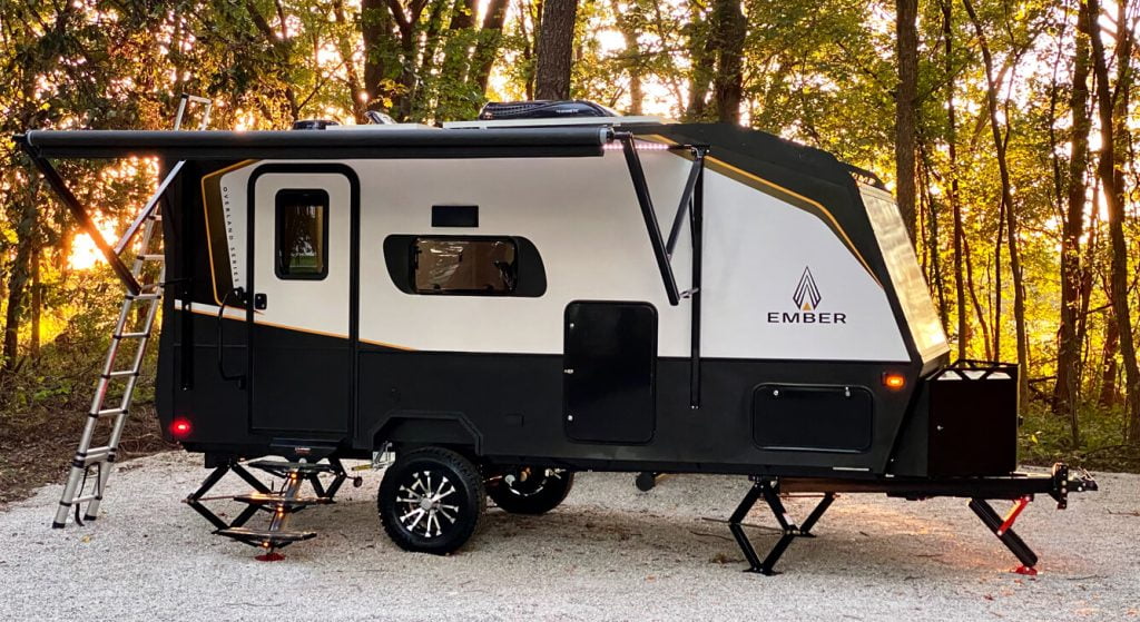 An Ember overland trailer, parked on a flat campsite surrounded by trees. Beyond the trees, the sun is setting.