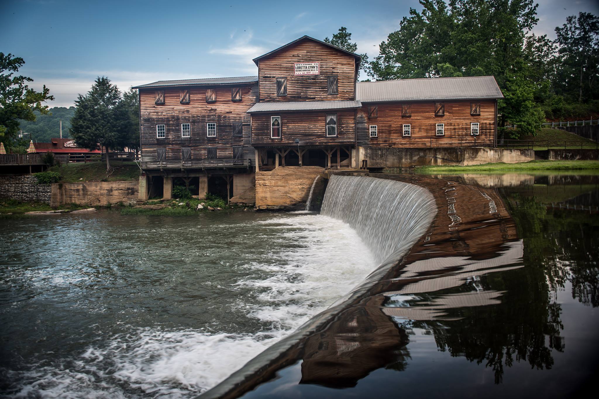 The exterior of a historic grist mill. The two-story building is clad in natural wood siding, darkened and weathered in spots. The mill backs onto the running waters of Hurricane Creek. 