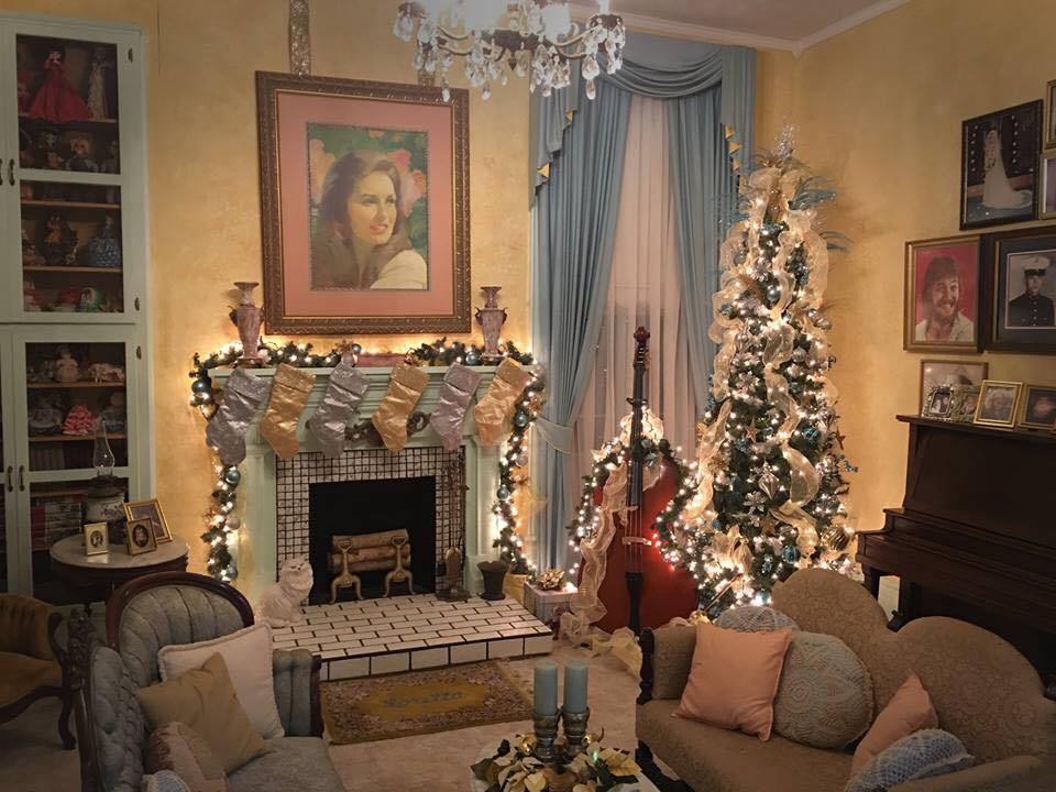 The sitting room of Loretta Lynn’s mansion, featuring high ceilings, crystal chandelier, and a tiled fireplace. The space is decorated with elegant vintage furniture, a double bass, and piano adorned with family photos. The room is decorated for the holidays with a Christmas tree, lighted garland, and six stockings hanging from the mantle. 