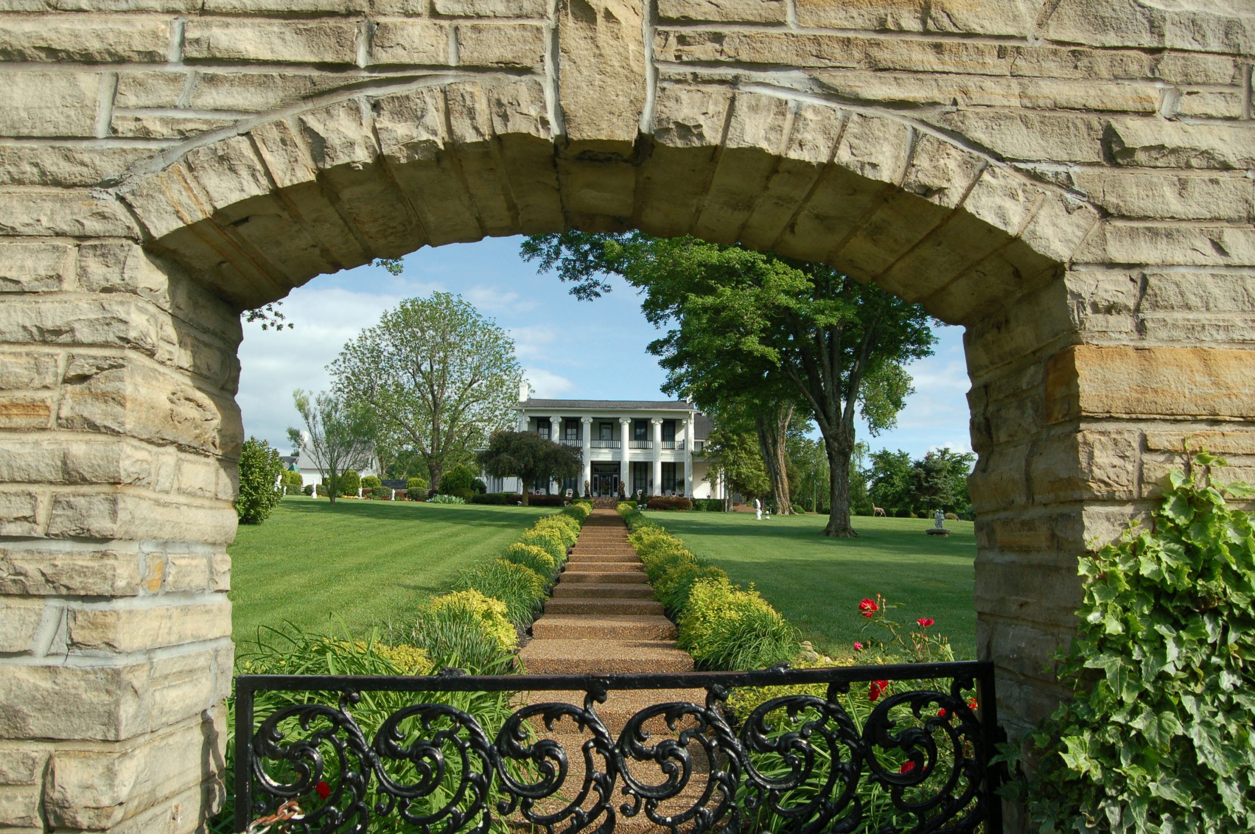 Loretta Lynn’s Ranch, main house – lush green lawns and a stepped walkway lead up to the big two-storey white house embellished with pillars – as viewed from a distance through a brick archway.