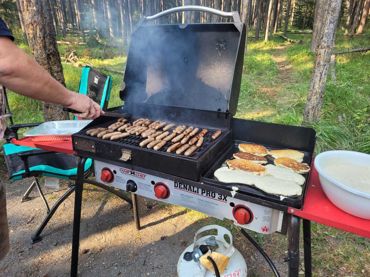 A giant breakfast being prepared on a camping barbeque. The grill is filled with sausages, browning. On the griddle, eight large pancakes are cooking. Keyword: how to plan for meals while RVing.