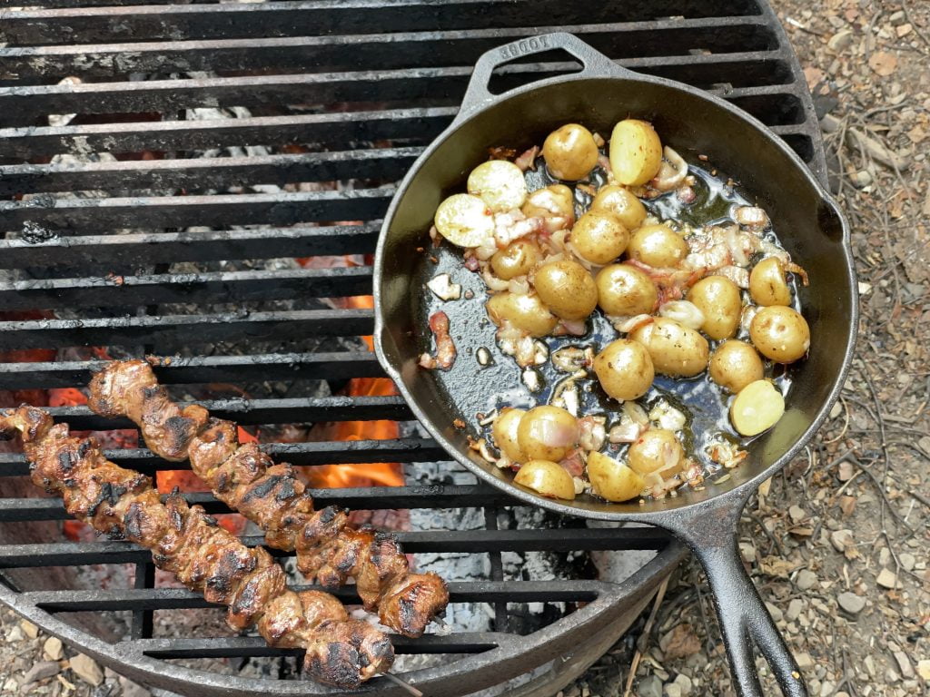 Two meat skewers and a cast iron frying pan filled with diced potato, garlic, and bacon, roasting over an open campfire.