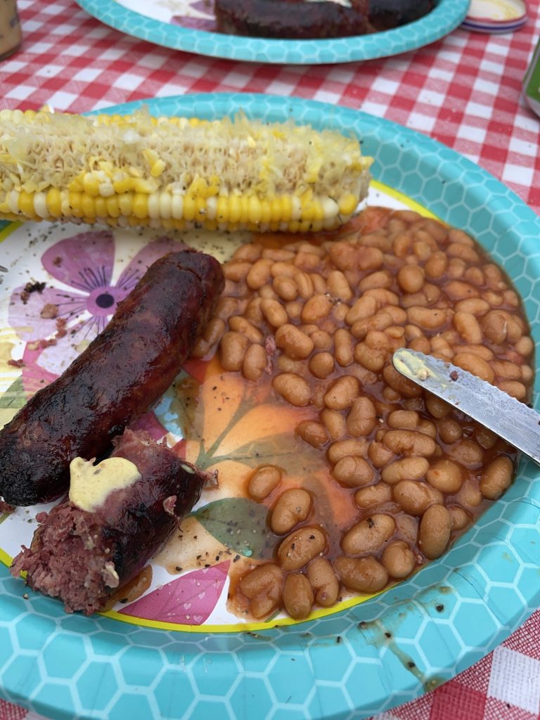 A disposable plate sitting on a red checkered tablecloth, the plate is filled with grilled sausage, baked beans, and corn on the cobb.