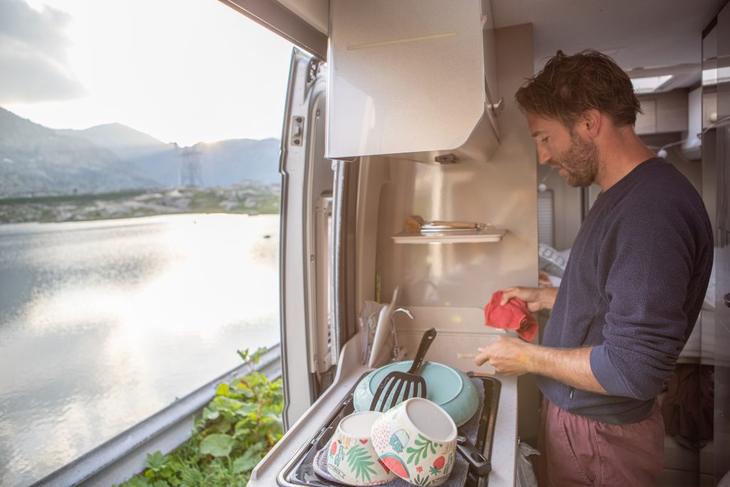 A white man in his early 40s, with bedhead, is standing at the sink in his RV washing dishes. Through the window over the sink, you can see a lake with a mountain range in the distance.