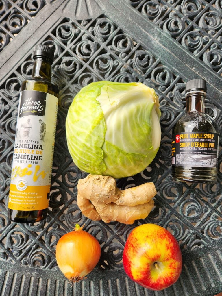 A bottle of camelina oil, cabbage, maple syrup, ginger, an onion, and an apple are displayed on a table.