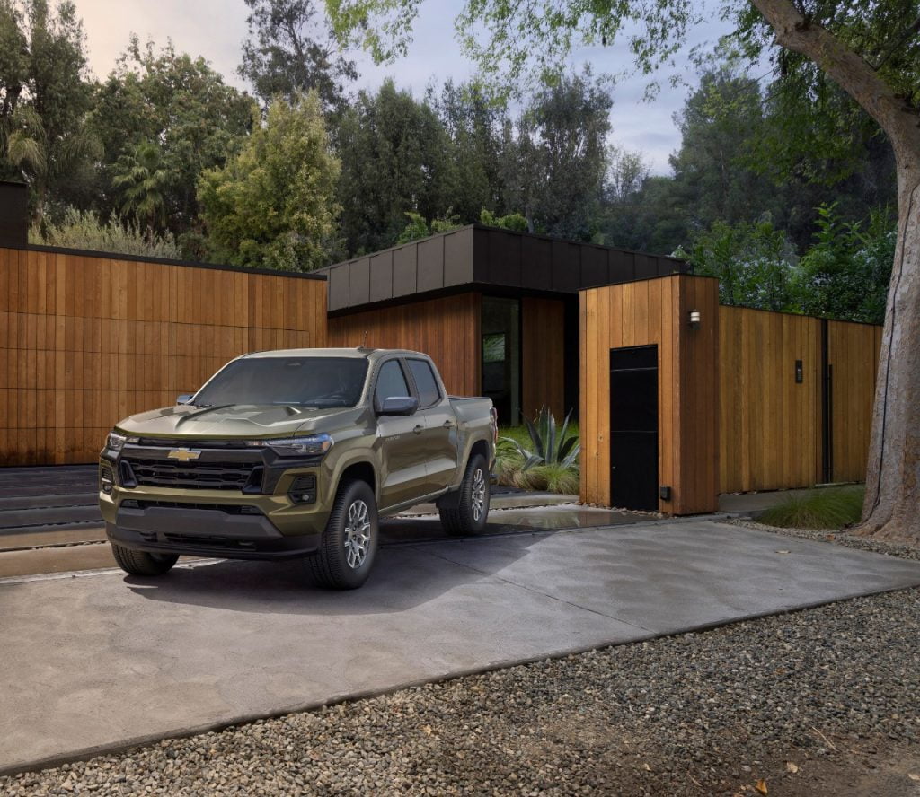 An olive green 2023 Chevrolet Colorado parked in the driveway of a contemporary home with vertical wood siding and black trim.