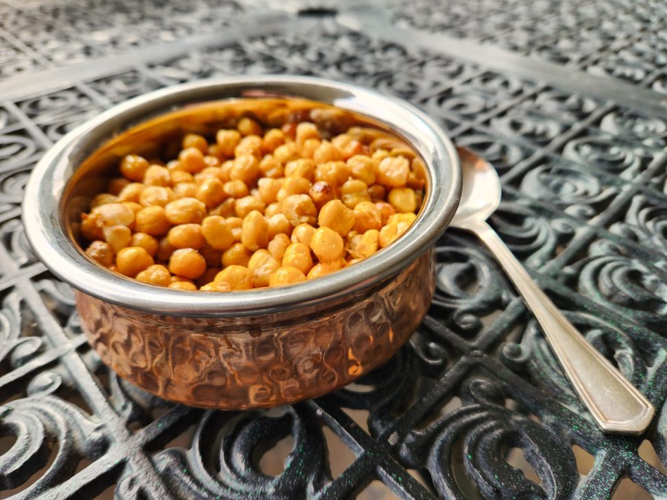A hammered copper bowl of roasted chickpeas with a silver spoon beside it.