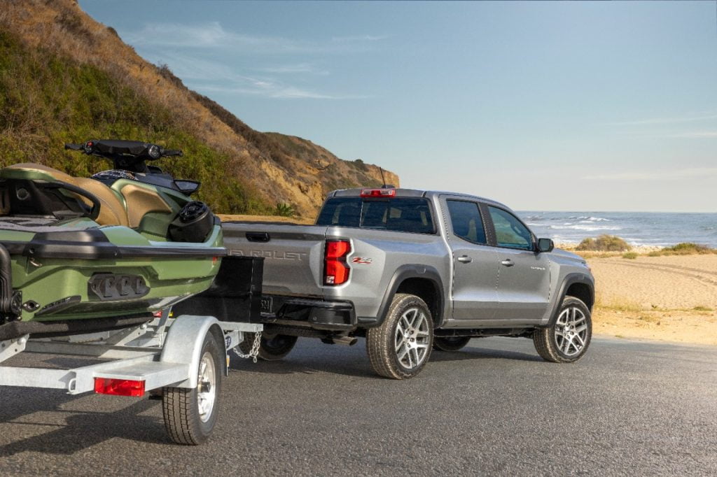 A silver 2023 Chevrolet Colorado pulling a Jet Ski down a paved road toward the beach.
