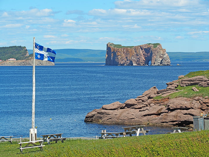 A photo, taken from the shoreline at Bonaventure Island looking out at the water. In the foreground, several picnic tables are set up on the grass while the Quebec provincial flag blows in the wind atop a wooden flagpole.
