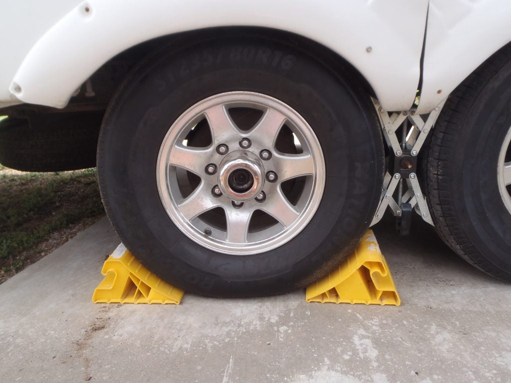 A close up of a camper trailer wheel, parked on a concrete pad. There is a yellow plastic wedge-shaped block on either side of the wheel to keep the trailer from rolling. 